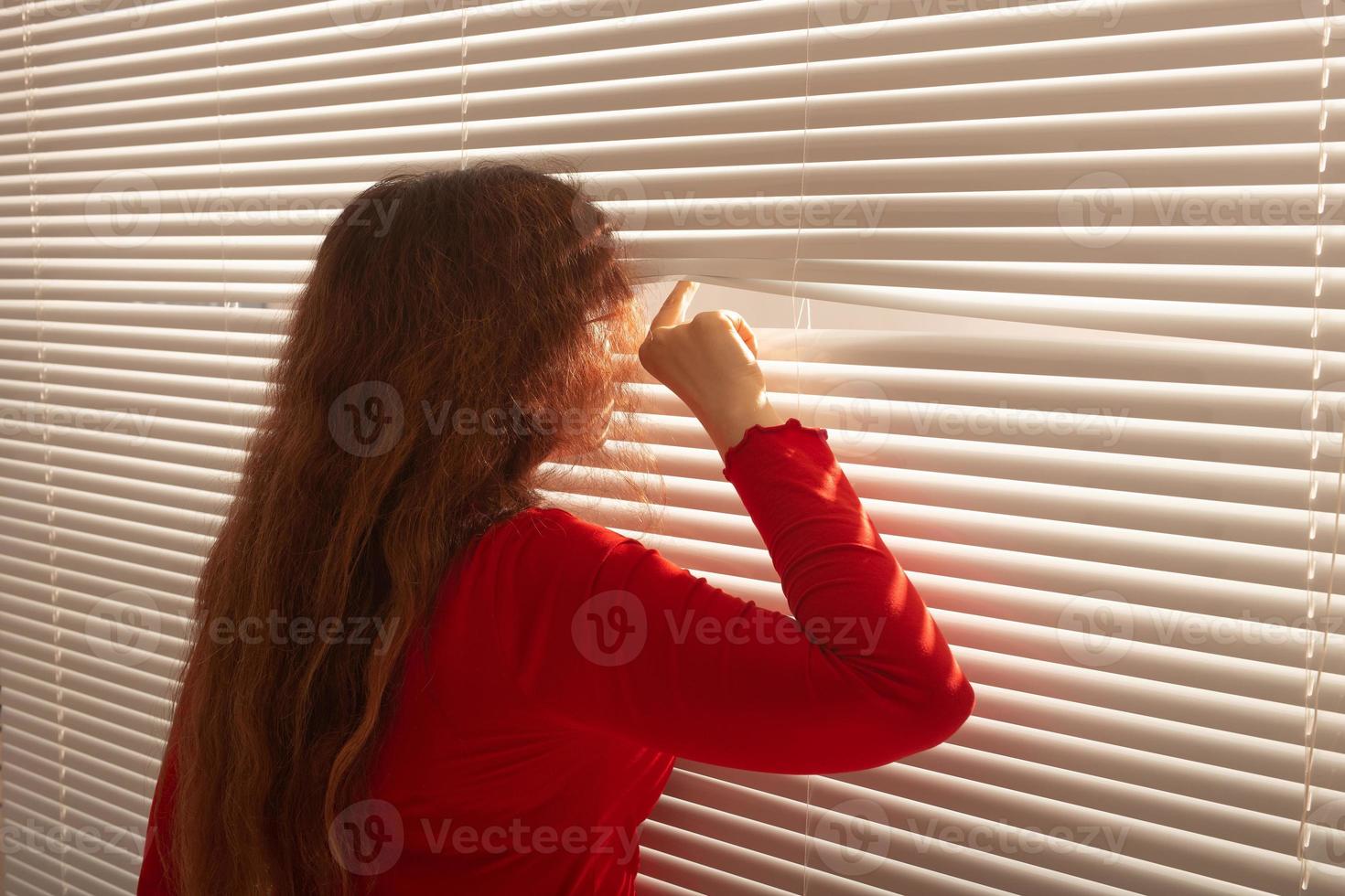 achterzijde visie van mooi jong vrouw met lang haar- gluurt door gat in de venster jaloezieën en looks uit de venster. toezicht en nieuwsgierigheid concept foto