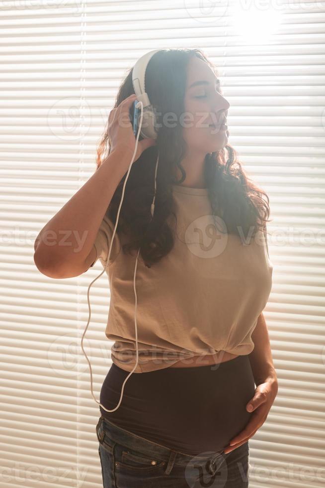 krullend haar brunette gepacificeerd zwanger vrouw luistert naar aangenaam klassiek muziek- gebruik makend van smartphone en koptelefoon. concept van een rustgevend humeur voordat vergadering baby. foto