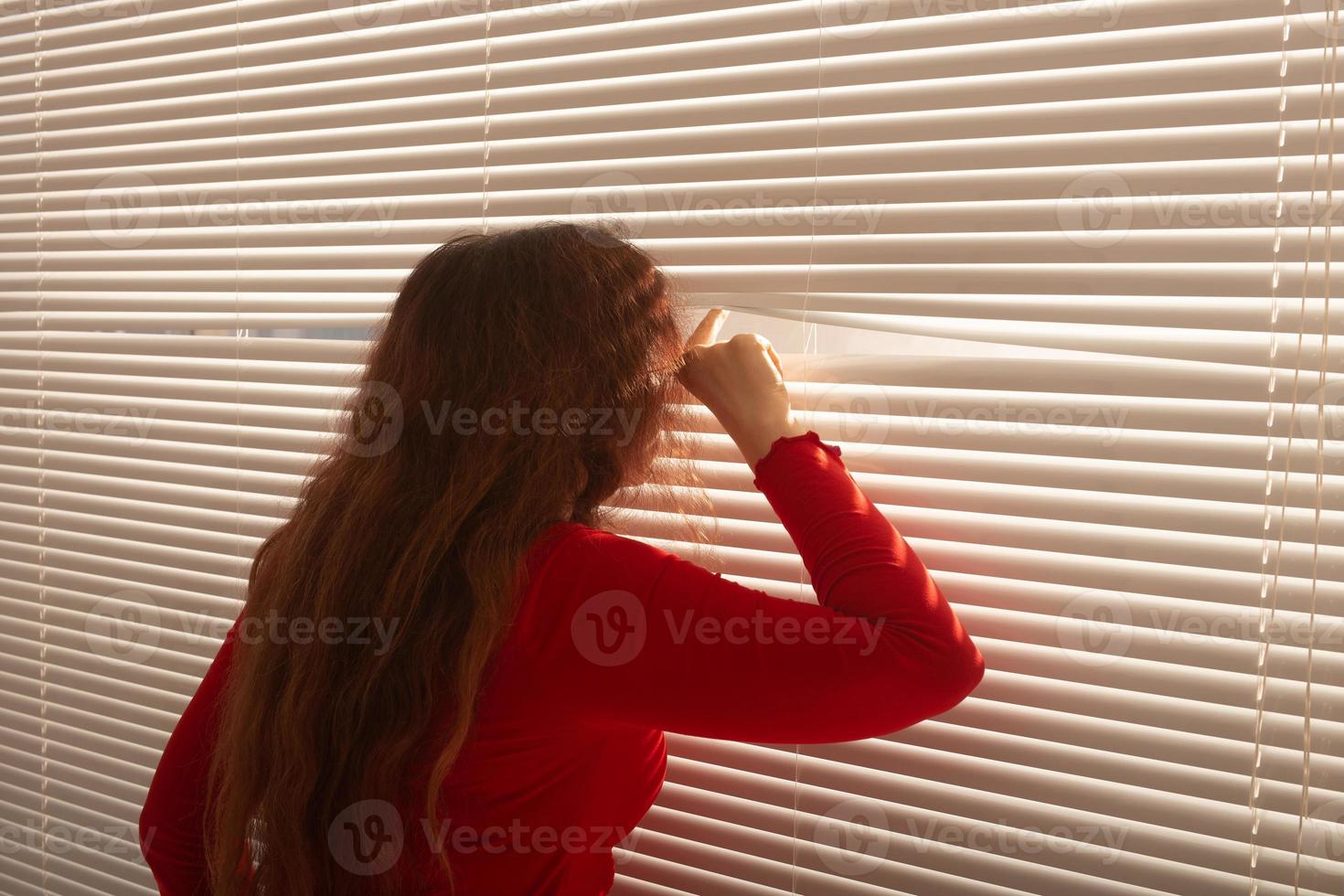 achterzijde visie van mooi jong vrouw met lang haar- gluurt door gat in de venster jaloezieën en looks uit de venster. toezicht en nieuwsgierigheid concept foto