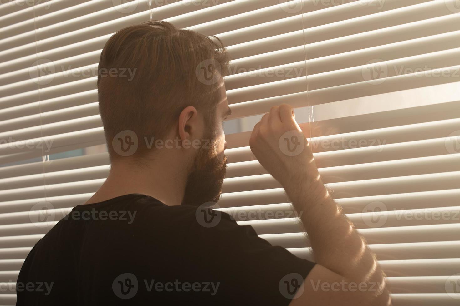 achterzijde visie van jong Mens met baard gluurt door gat in de venster jaloezieën en looks uit in de straat. toezicht en nieuwsgierigheid concept foto