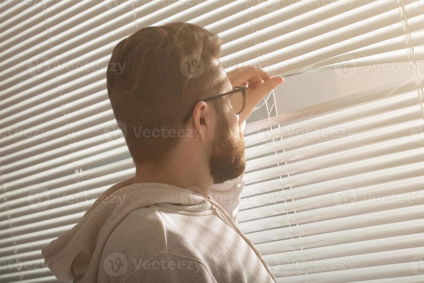 achterzijde visie van jong Mens met baard gluurt door gat in de venster jaloezieën en looks uit in de straat. toezicht en nieuwsgierigheid concept foto