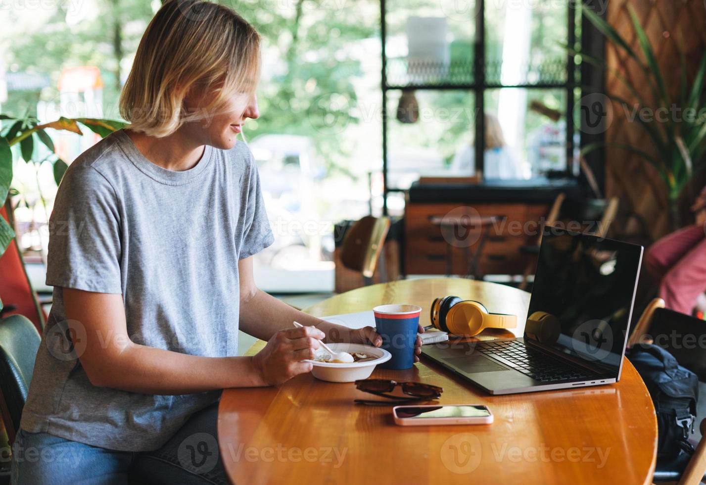 jong glimlachen blond vrouw freelancer met geel hoofdtelefoons werken Aan notitieboekje Aan tafel Bij cafe. meisje leerling meisje aan het studeren Aan online Cursus. vrouw hebben ontbijt in de cafe foto