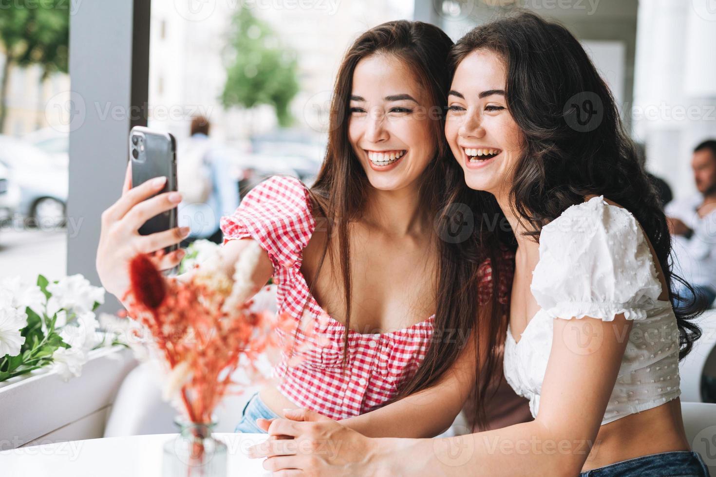 gelukkig brunette jong meisjes vrienden in zomer kleren nemen selfie Aan mobiel telefoon in de zomer straat cafe foto