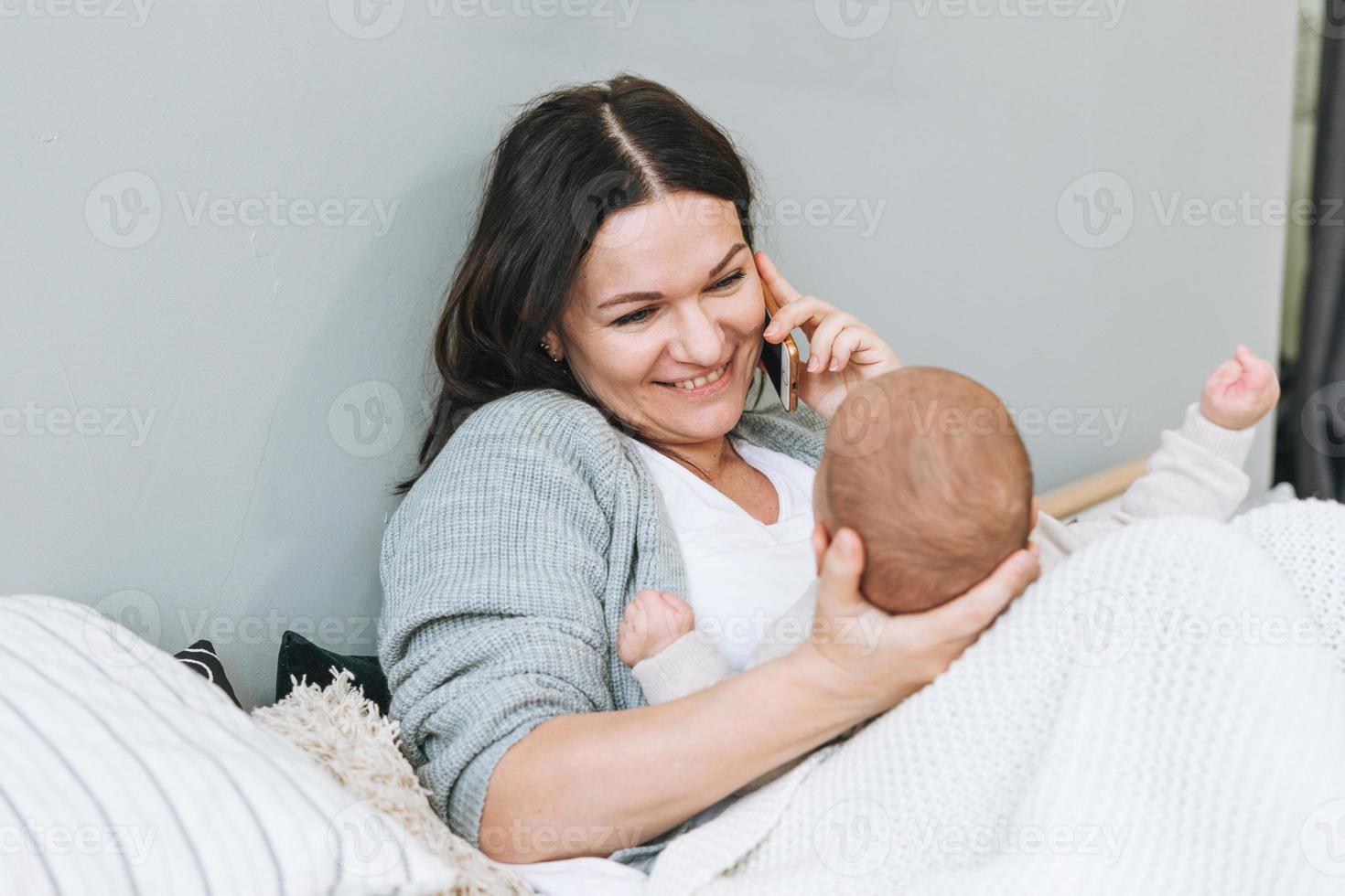 moeder sprekend Aan mobiel telefoon met schattig baby jongen Aan de bed, natuurlijk tonen, liefde emotie foto