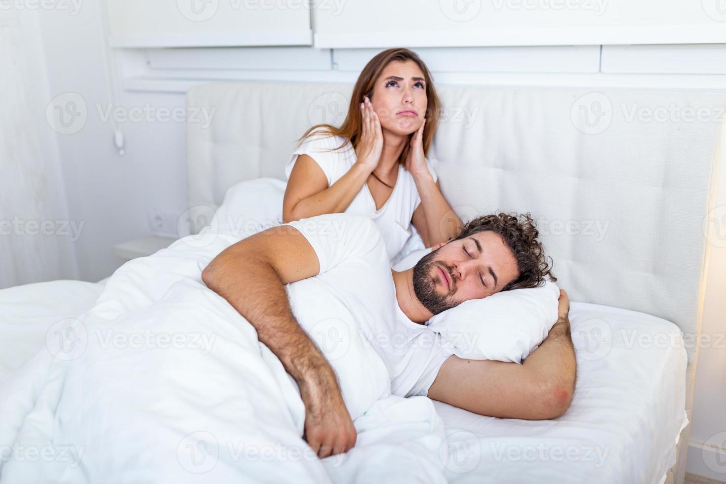 Mens snurken terwijl zijn vrouw is aan het bedekken oren met handen. vrouw lijdt van haar mannetje partner snurken in bed. lawaai concept. echt mensen. jong geërgerd vrouw aan het liegen in bed met snurken man foto