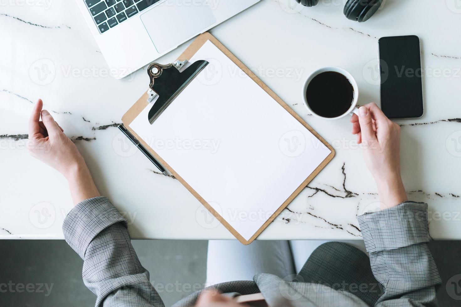 top visie van bedrijf vrouw Aan werken plaats met kop van koffie, notities, Open laptop, smartphone Aan wit marmeren tafel in de helder modern kantoor kamer, mockup foto