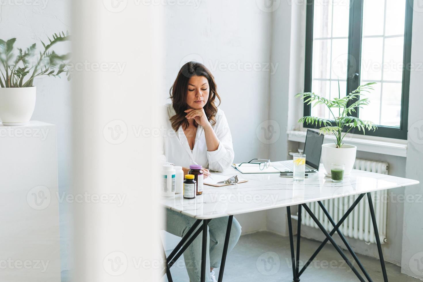 jong glimlachen brunette vrouw dokter voedingsdeskundige plus grootte in wit overhemd werken Bij laptop Bij modern helder kantoor kamer. de dokter schrijft voor een voorschrift voor geneesmiddelen en vitamines Bij kliniek foto