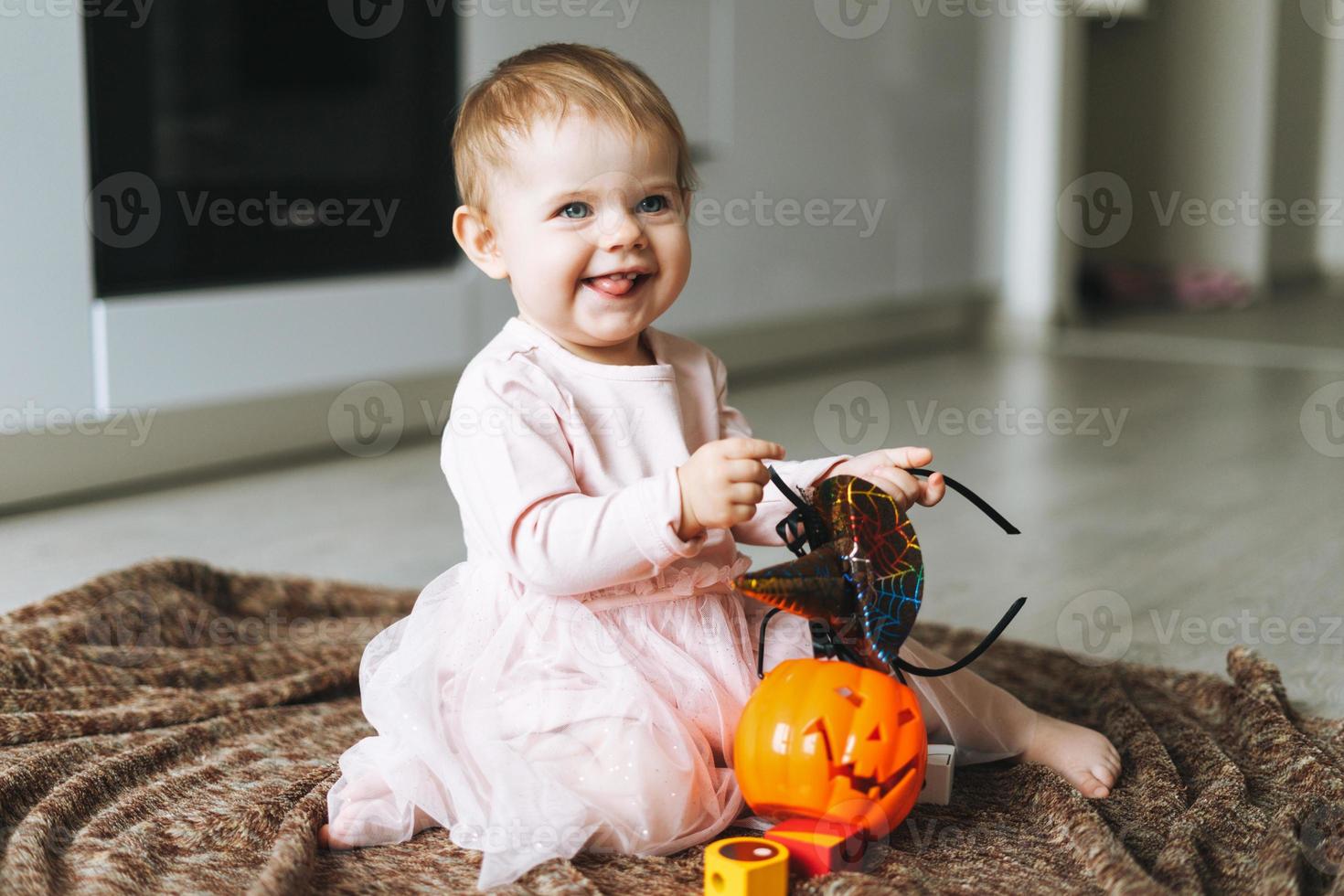 schattig weinig baby meisje in roze jurk met de heks hoed en pompoen lantaarn zittend Aan verdieping in keuken Bij de huis, halloween tijd foto