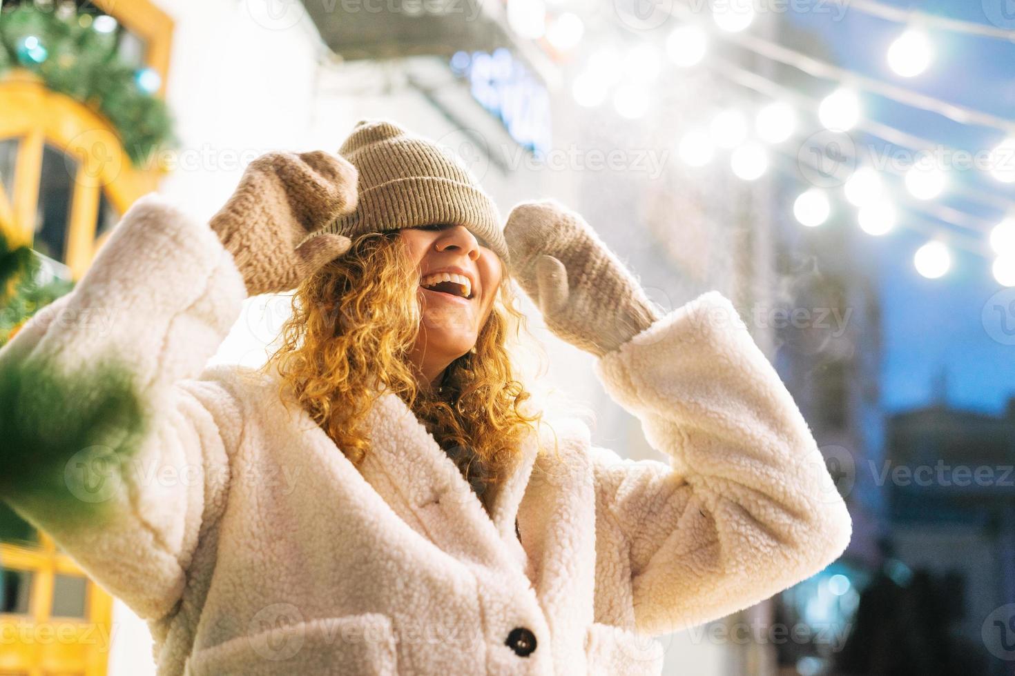 portret van jong gelukkig vrouw met gekruld haar- in vacht jasje hebben pret in winter straat versierd met lichten foto