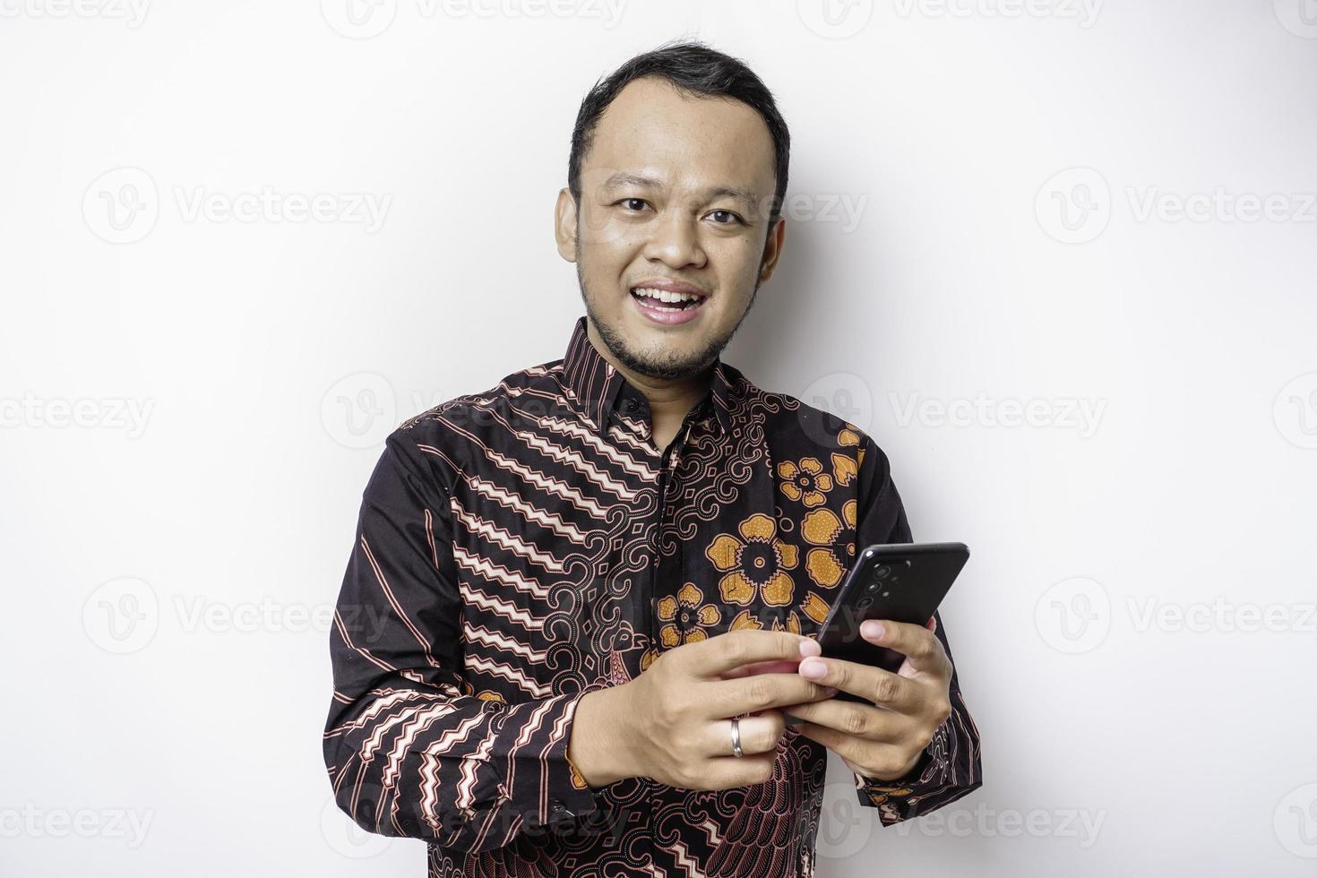 een portret van een gelukkig Aziatisch Mens vervelend batik overhemd en Holding zijn telefoon, geïsoleerd door wit achtergrond foto