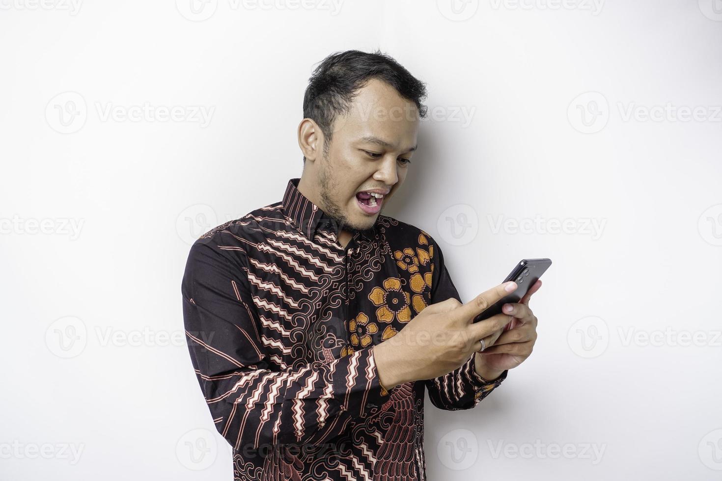 een portret van een gelukkig Aziatisch Mens vervelend batik overhemd en Holding zijn telefoon, geïsoleerd door wit achtergrond foto