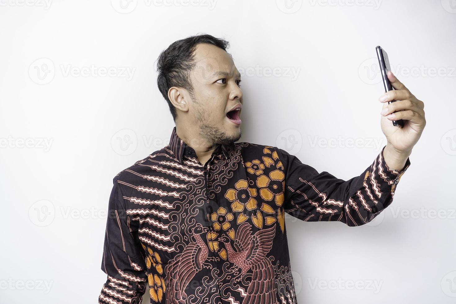 geschokt Aziatisch Mens vervelend batik overhemd en Holding zijn telefoon, geïsoleerd door wit achtergrond foto