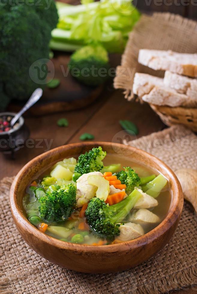 kip soep met broccoli, groen erwten, wortels en selderij in kom Aan een houten achtergrond in rustiek stijl foto