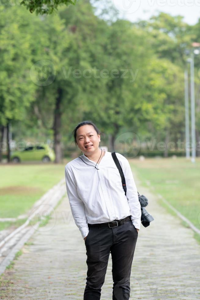 Aziatisch freelance lang haar- Mens staand en posten Aan de wandelen manier in de tuin buitenshuis veld- met zijn camera naast hem. foto