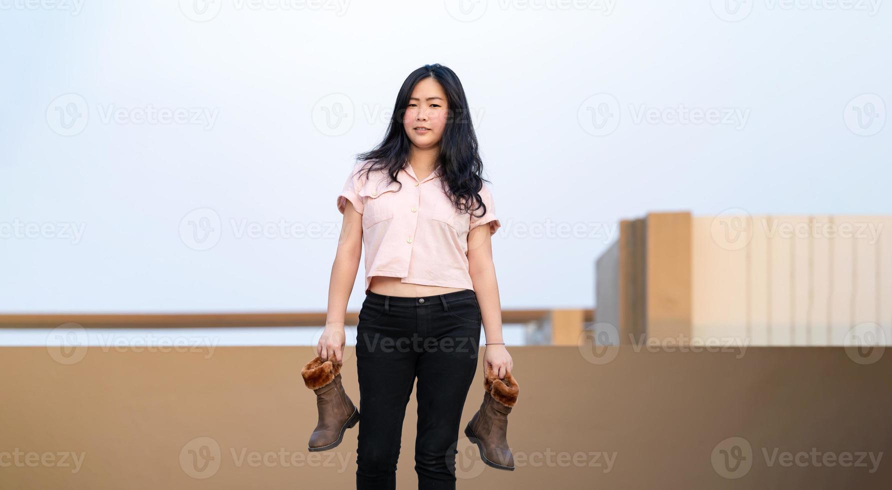 Aziatisch vrouw houdt bruin laarzen in haar hand- en posten voor foto het schieten Bij op het dak milieu Bij schemering tijd.