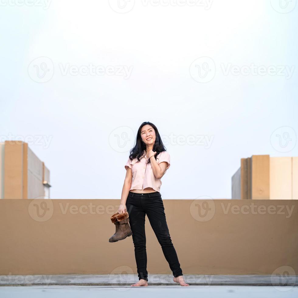 Aziatisch vrouw staat en glimlacht alleen met houdt laarzen Aan op het dak in pastel blauw - bruin schaduw. foto