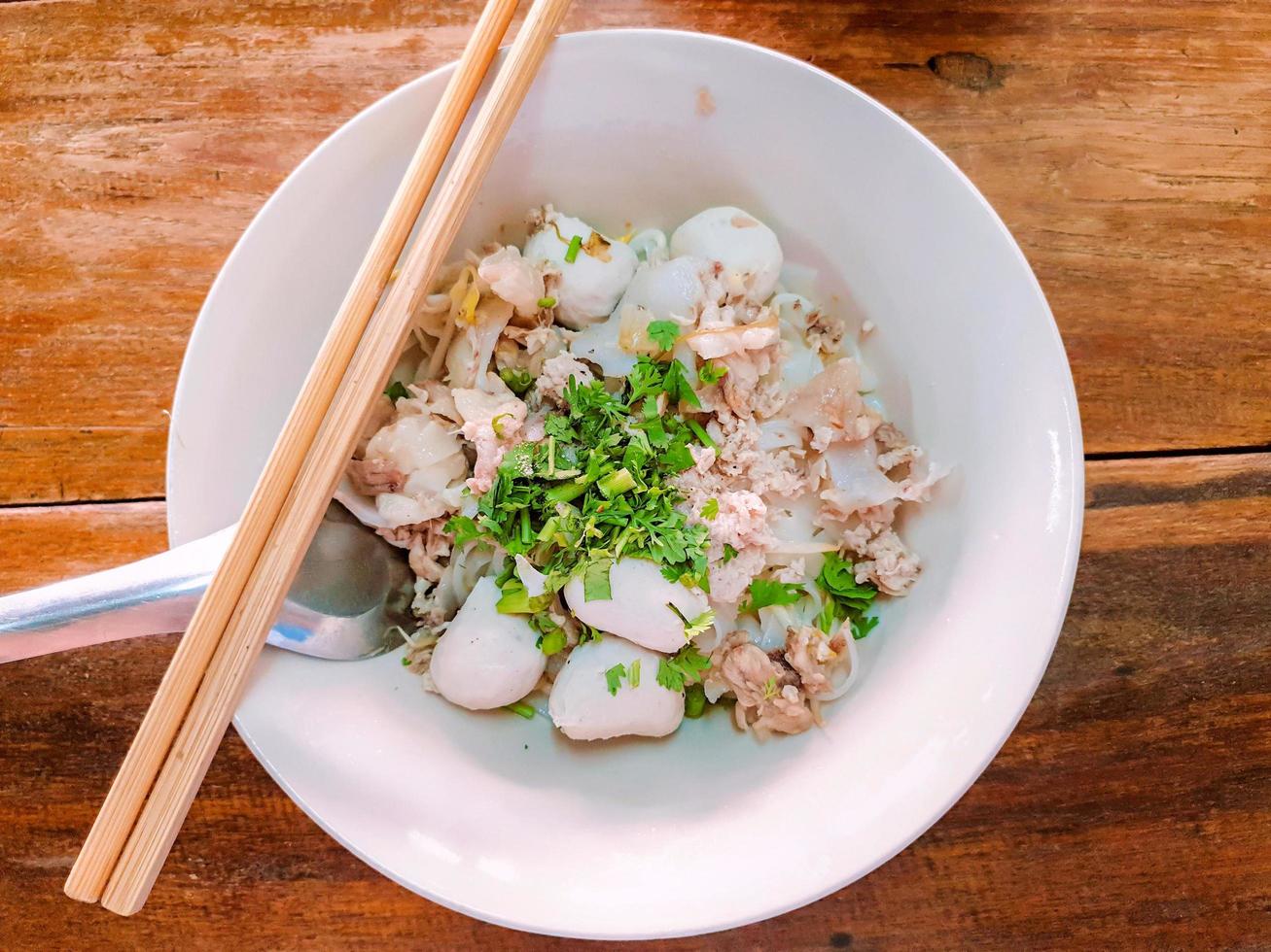 top visie en detailopname van lekker niet soep Chinese noodle met varkensvlees ballen en stuk van ongeveer varkensvlees in wit keramisch kom met eetstokjes en lepel Aan houten tafel achtergrond.. foto