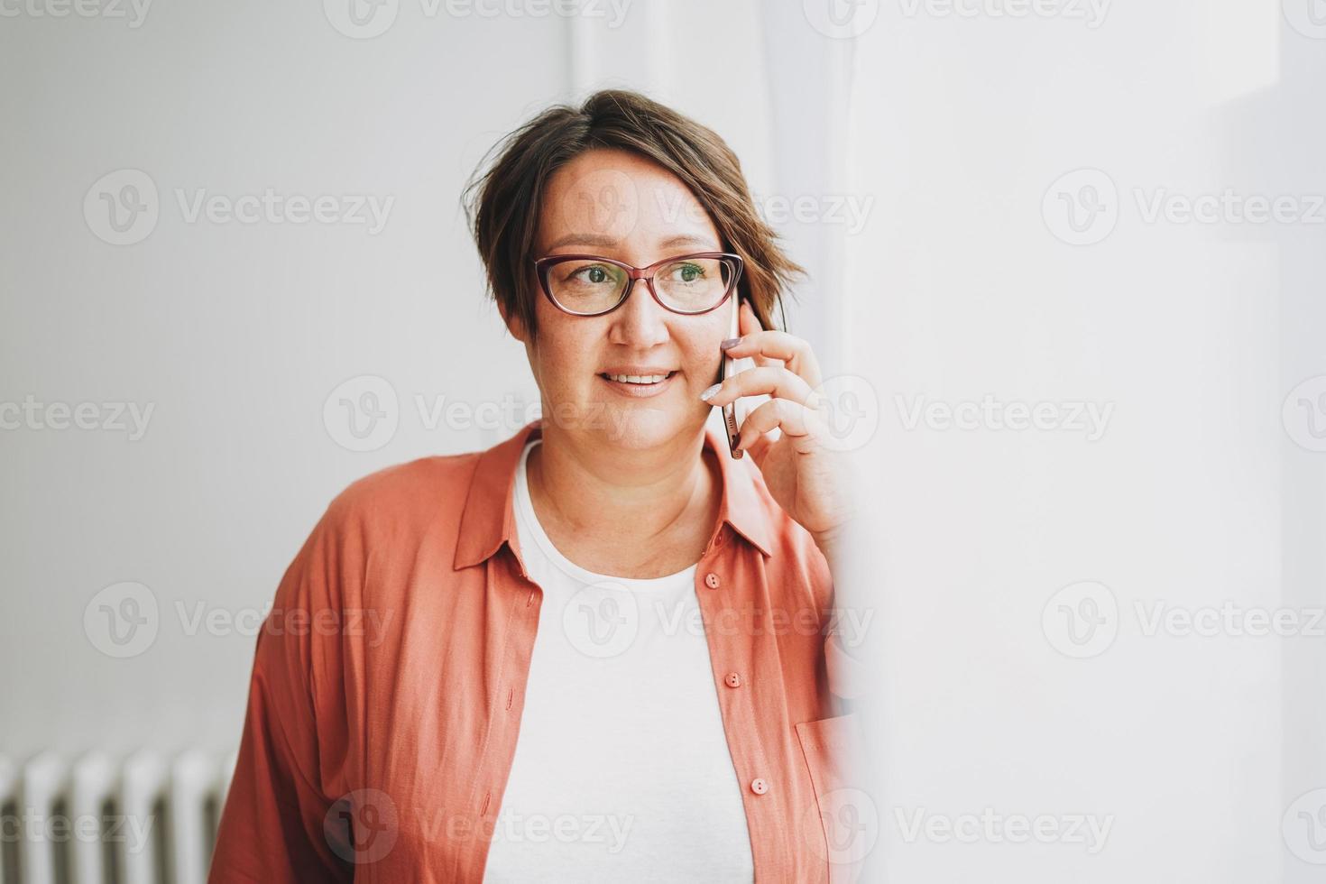 volwassen charmant brunette vrouw zakenvrouw in bril plus grootte lichaam positief gebruik makend van mobiel in de buurt venster foto