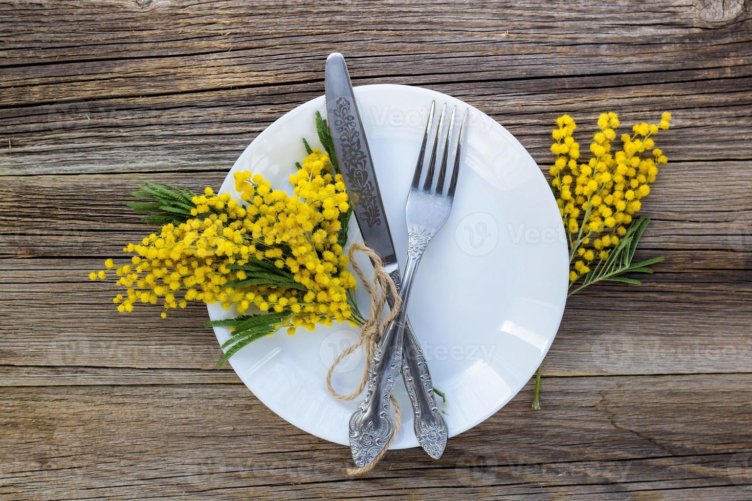 vork mes met bord en mimosa bloemen Aan houten tafel voor voorjaar Pasen vakantie diner. foto