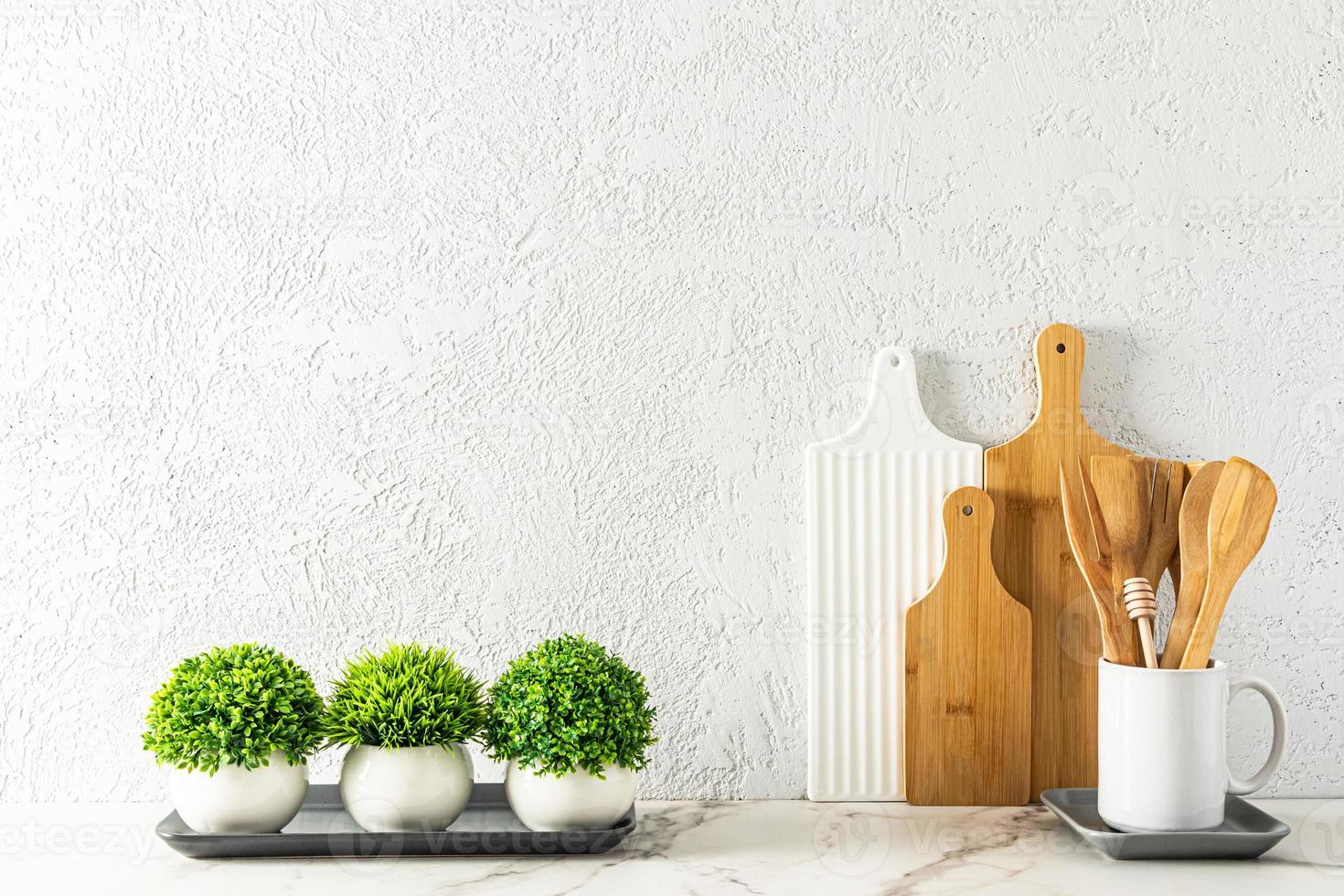 snijdend borden en houten lepels in een wit mok Aan een marmeren wit aanrecht tegen een grijs cement muur met groen kamerplanten. foto