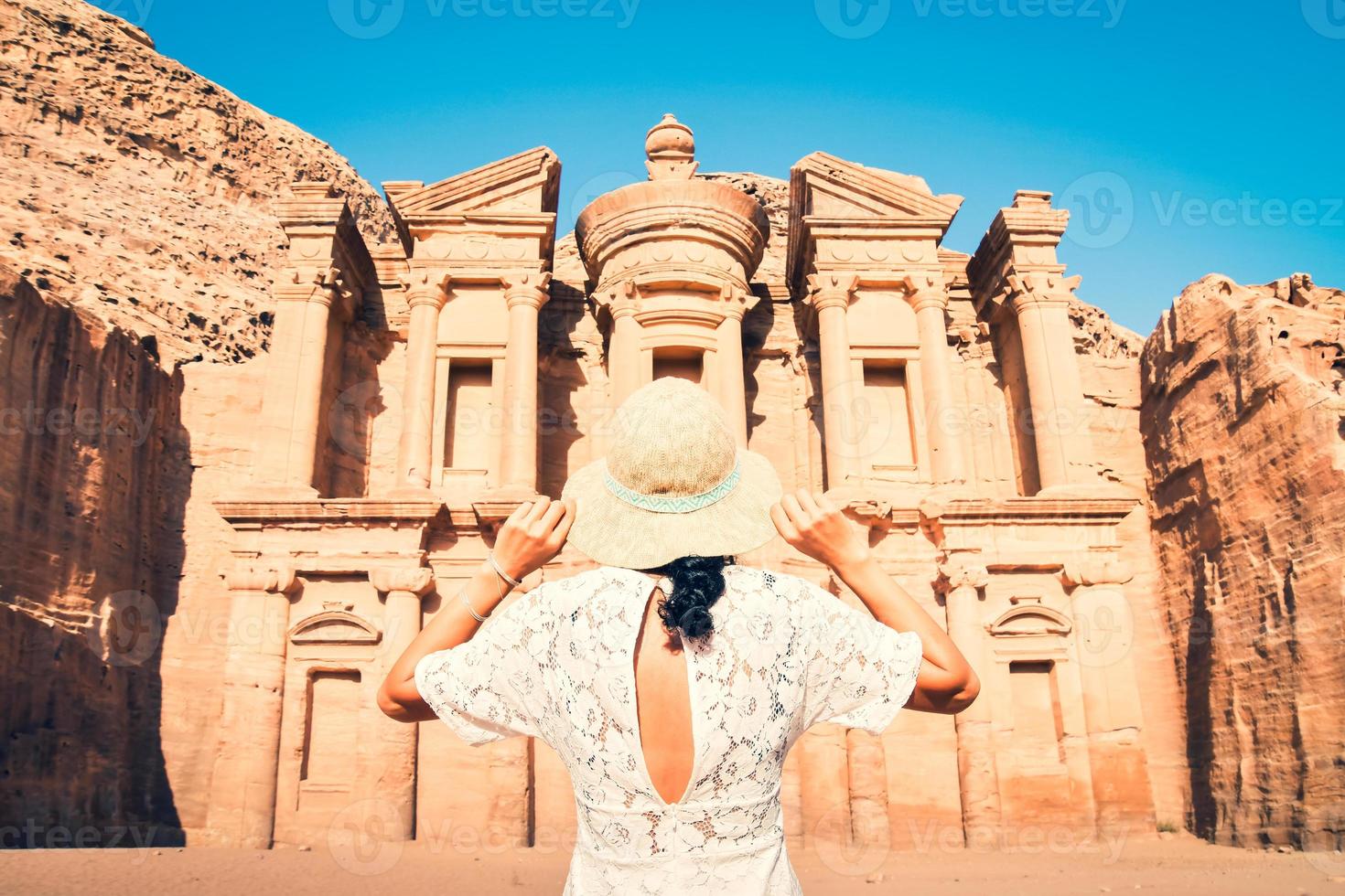 Kaukasisch vrouw toerist in wit jurk houden haar wit hoed houding Bij advertentie deir of el hun, de monument gesneden uit van rots in de oude stad petra, Jordanië. UNESCO wereld erfgoed plaats foto
