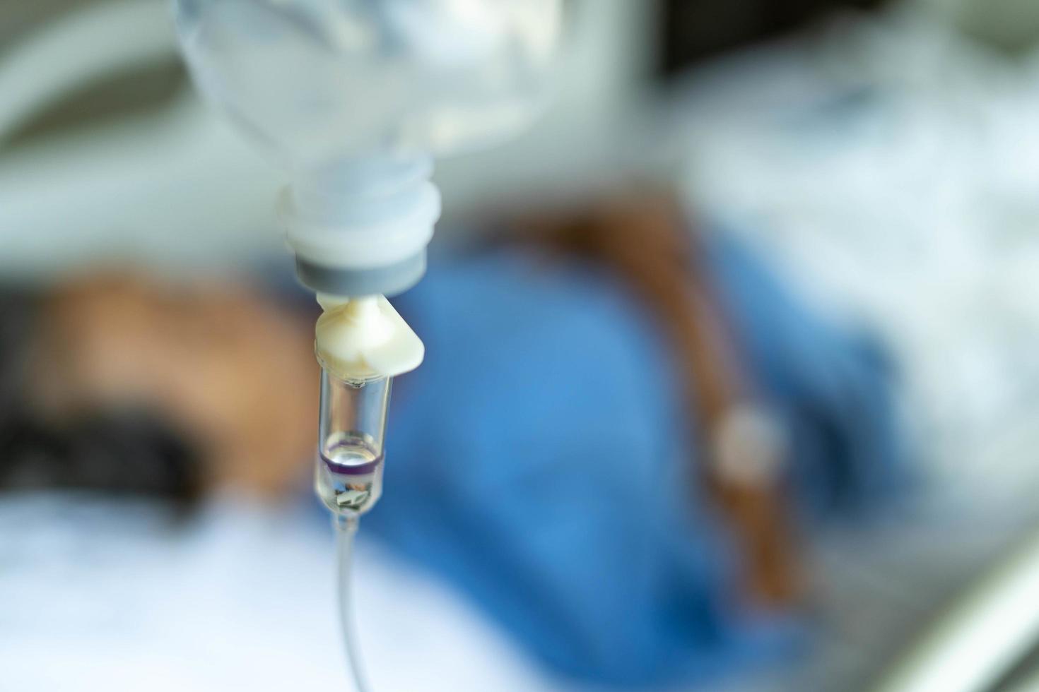 een detailopname van een zoutoplossing fles vastgebonden naar een naald- geprikt Bij de hand- van een vrouw geduldig aan het liegen in een ziekenhuis bed. medisch en behandeling van patiënten concepten foto