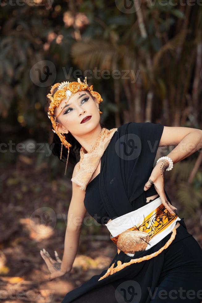 Aziatisch vrouw poses met haar hand- terwijl vervelend een zwart jurk en een goud riem met een goud kroon Aan haar hoofd foto