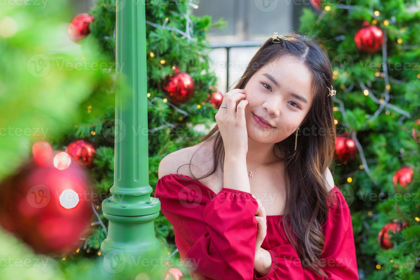 schattig Aziatisch vrouw in rood jurk staat glimlachen gelukkig op zoek Bij de camera in voorkant van de Kerstmis boom met bokeh net zo achtergrond in de thema van Kerstmis en nieuw jaar vieringen foto