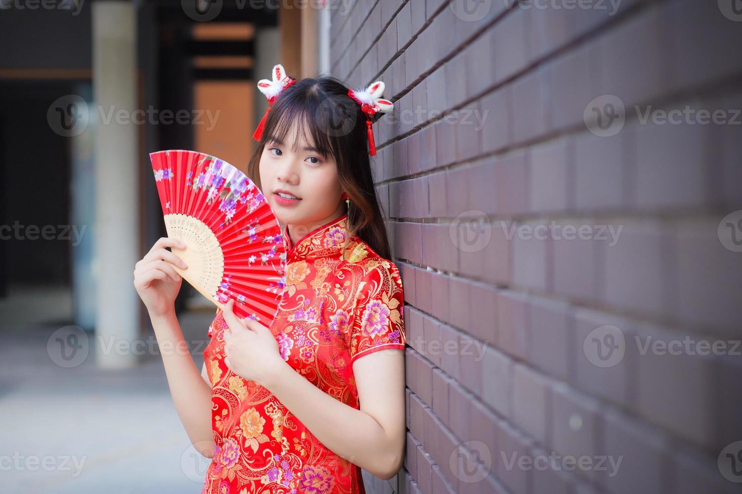 mooi Aziatisch vrouw in rood jurk staat glimlachen gelukkig op zoek Bij de camera houdt een ventilator tussen oud stad centrum in Chinese nieuw jaar thema. foto