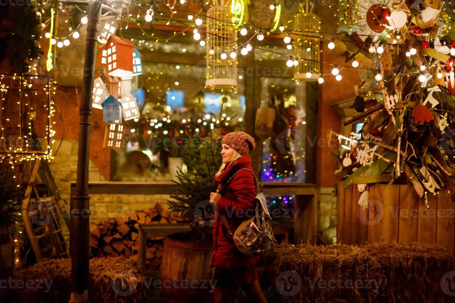 buitenshuis foto van jong mooi gelukkig glimlachen meisje poseren in straat. feestelijk Kerstmis eerlijk Aan achtergrond. model- vervelend elegant winter jas, gebreid muts hoed, sjaal.