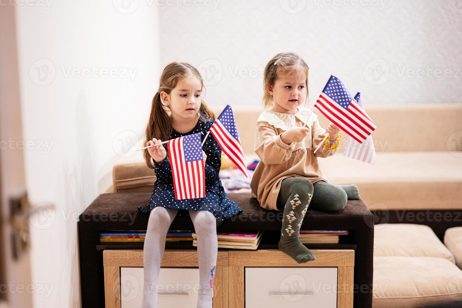 twee zussen zijn zittend Aan een bankstel Bij huis met Amerikaans vlaggen Aan handen. Verenigde Staten van Amerika kinderen meisjes met vlag . foto