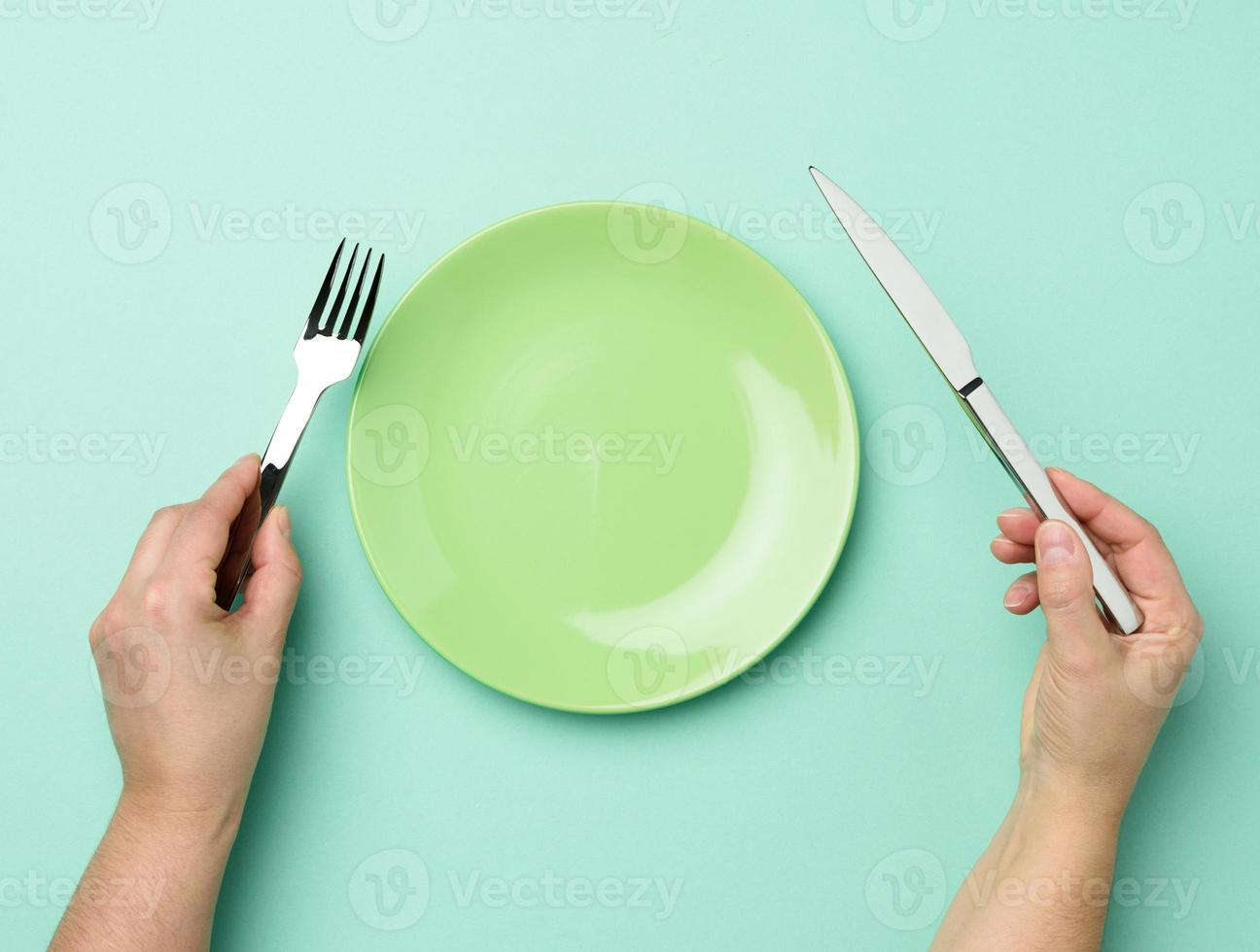 twee handen houden een metaal mes en vork Aan de oppervlakte van een ronde leeg groen bord foto