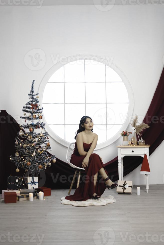 portret van mooi jong meisje knus zitten omlaag, glimlachen slijtage rood japon in versierd Kerstmis leven kamer binnenshuis foto