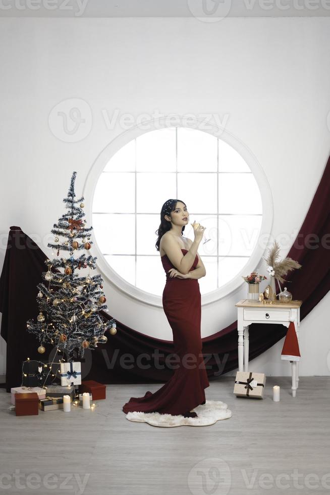 portret van een mooi jong meisje vervelend een rood japon en drinken wijn, glimlachen Bij de camera, staand in versierd Kerstmis leven kamer binnenshuis foto