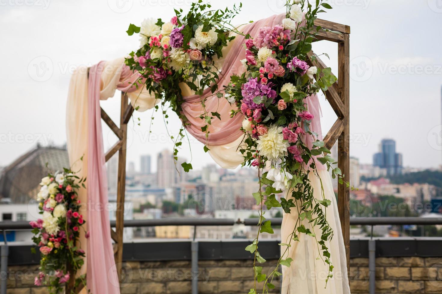 mooi bruiloft ceremonie buitenshuis. bruiloft boog gemaakt van kleding en wit en roze bloemen Aan de dak tegen de backdrop van een groot stad . oud deuren, rustiek stijl. foto