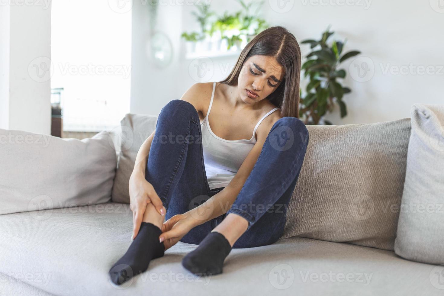 handen masseren gezwollen voet terwijl zittend Aan sofa gedurende de dag Bij huis. foto van jong Kaukasisch vrouw lijden van pijn in been. vrouw masseren haar poten na allemaal dag Bij werk in kantoor