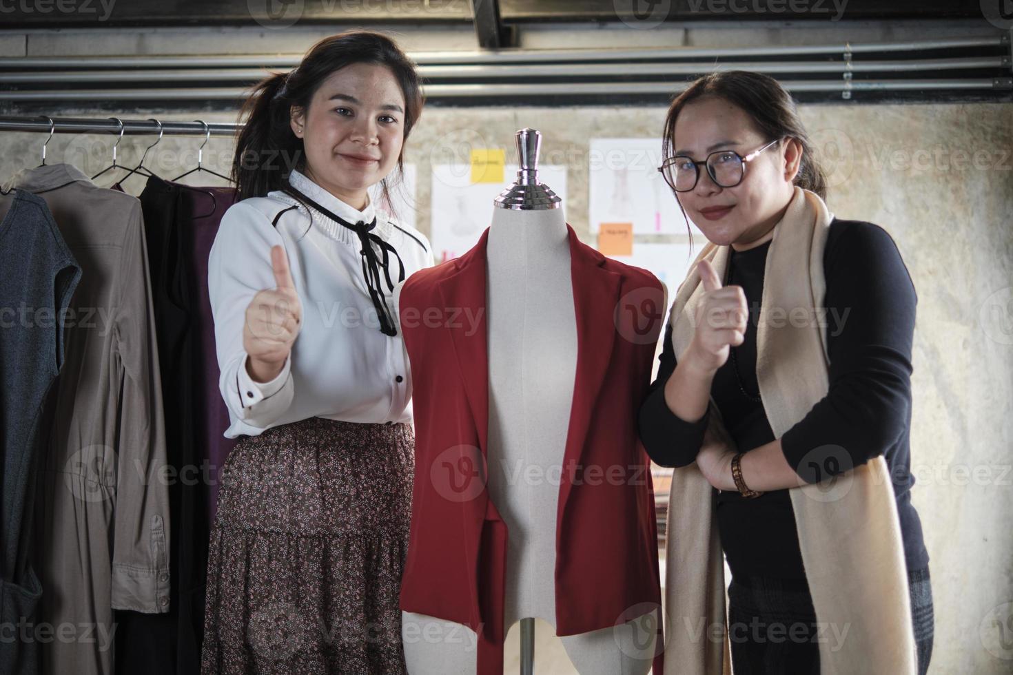 mode team mensen, Aziatisch vrouw ontwerper en tiener assistent in studio, duim omhoog en glimlach, gelukkig werken met draad en naaien voor jurk ontwerp, professioneel winkel kleermaker mkb ondernemer. foto