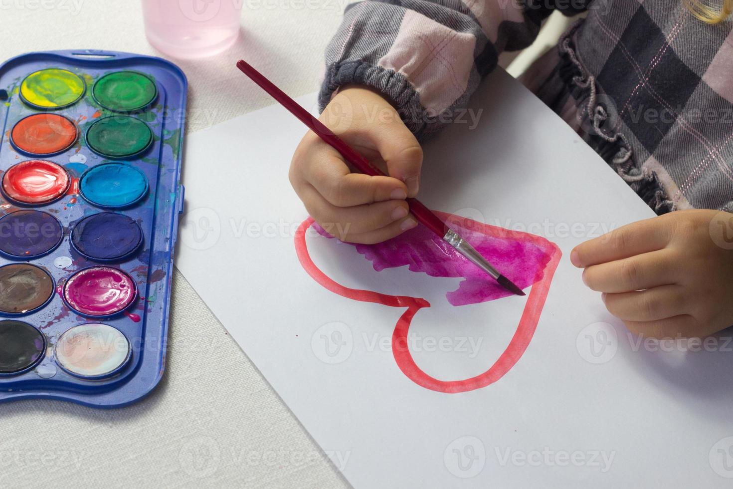 weinig meisje zittend Bij de tafel en tekening rood hart aquarel, valentijn dag ansichtkaart, groet kaart, kunst school- concept, reclame, kind schilderij waterverf Bij thuis, peuter activiteit, kinderen vrije tijd. foto