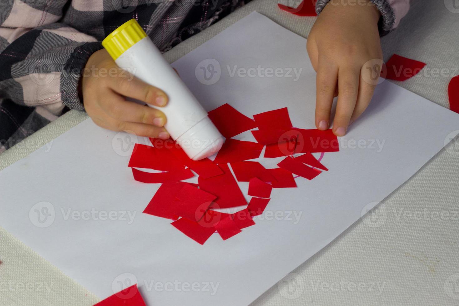 weinig meisje maken rood hart stoffen met papier en lijm Aan wit blad, valentijnsdag dag groet kaart, vakantie geschenk voor moeders dag, handen gemaakt kinderen heden, liefde symbool met papier, peuter werkzaamheid foto