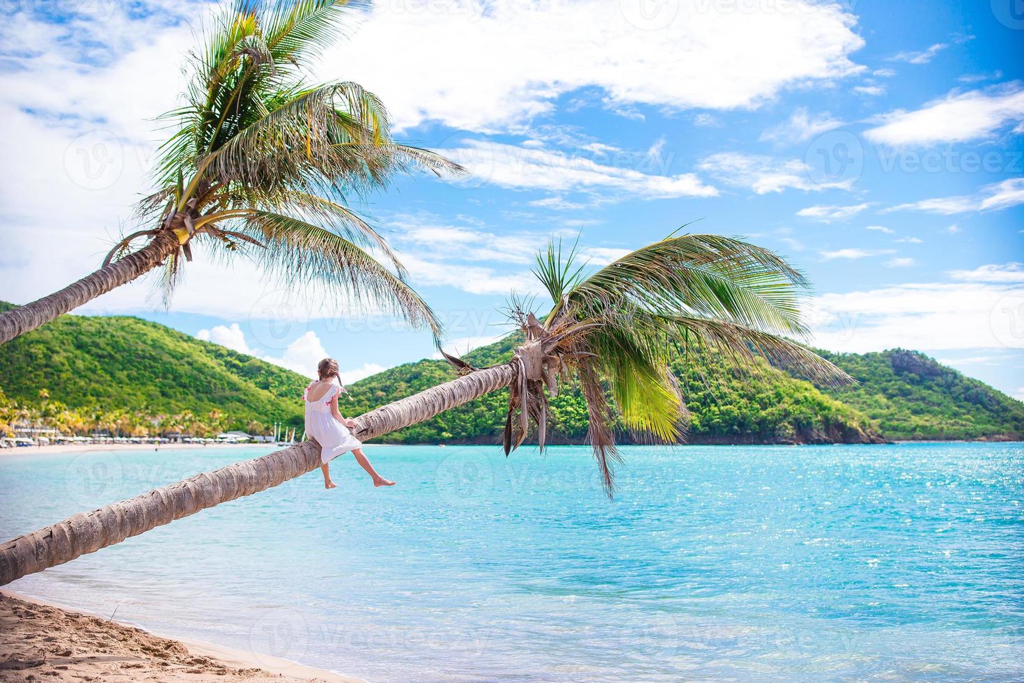 aanbiddelijk weinig meisje zittend Aan palm boom gedurende zomer vakantie Aan wit strand foto