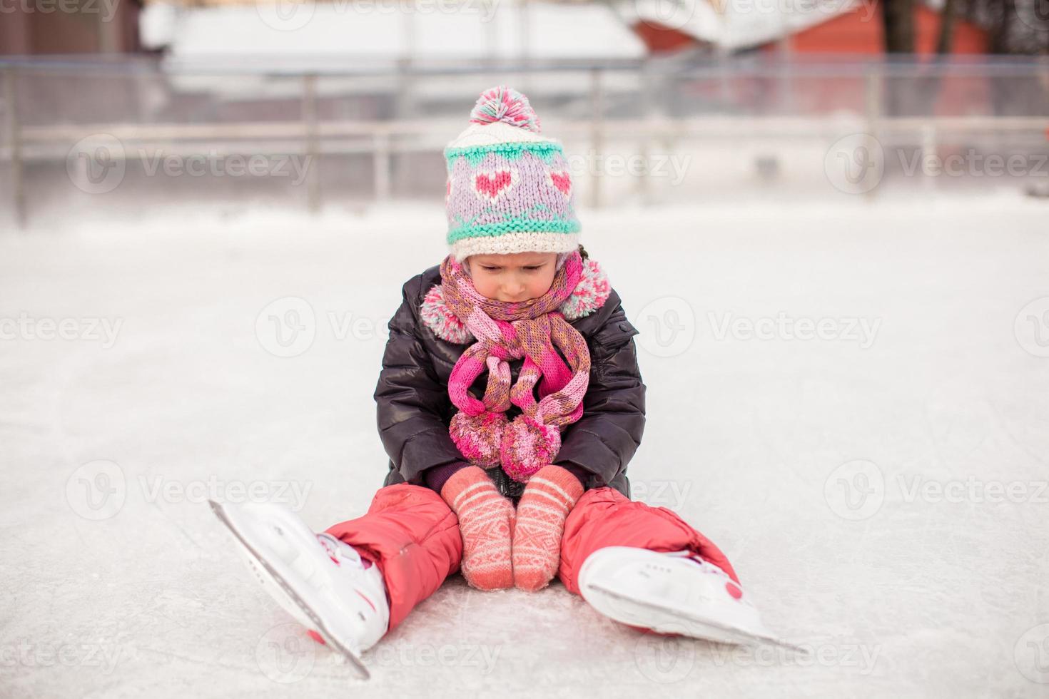 weinig verdrietig meisje zittend Aan een het schaatsen baan na de vallen foto