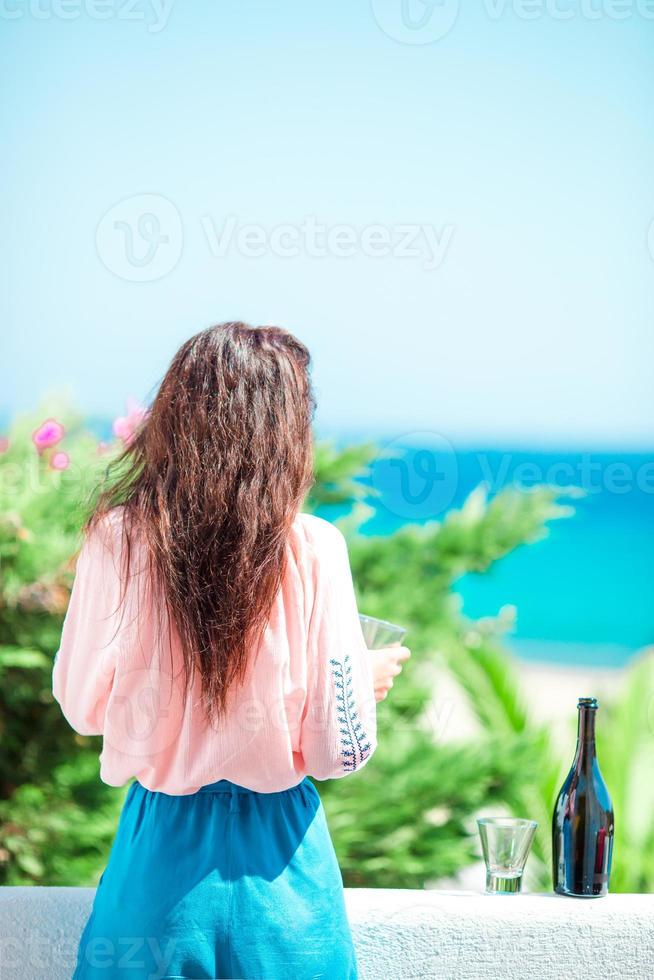 vrouw met bril en fles van smakelijk wijn Aan balkon in Grieks eiland met visie van zee foto