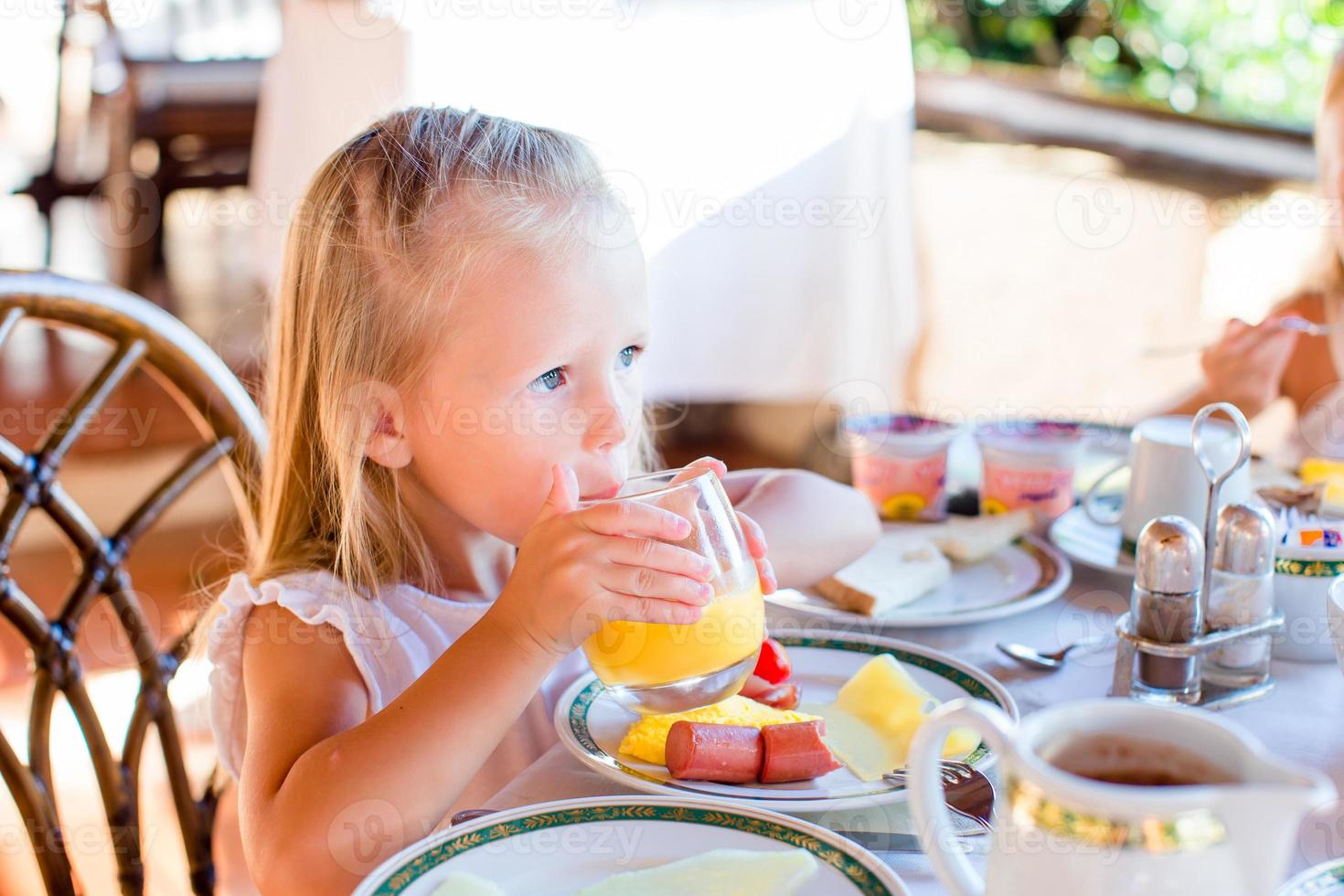 aanbiddelijk weinig meisje hebben ontbijt Bij restaurant. schattig kind genieten vers oranje sap in buitenshuis cafe foto