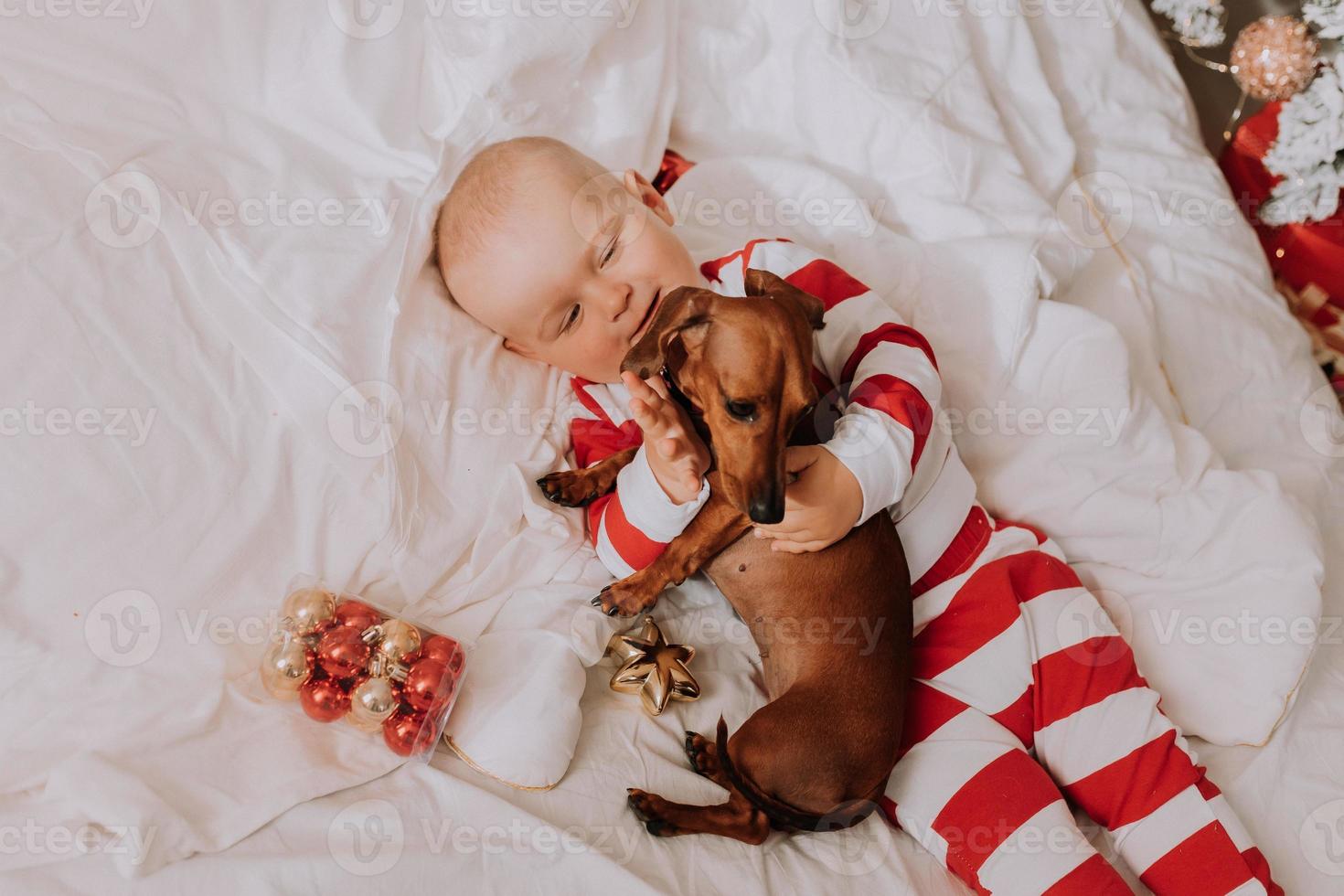 weinig jongen in rood en wit pyjama is aan het liegen in bed Aan een wit vel met een hond. Kerstmis ochtend. gezond slaap van de kind. ruimte voor tekst. hoog kwaliteit foto