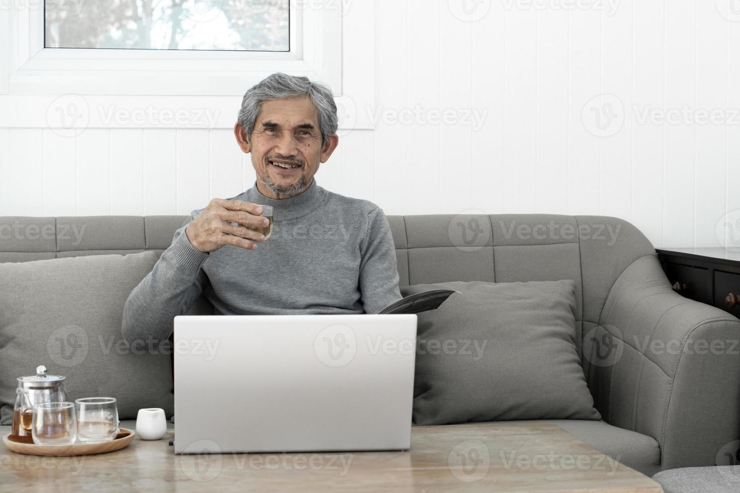 ouderen Aziatisch grijs haar- Mens in lang mouw overhemd zit Aan sofa en drinken heet thee in leven kamer van zijn huis, concept voor gelukkig leven van oudsten en leven verzekering in de omgeving van de wereld. foto