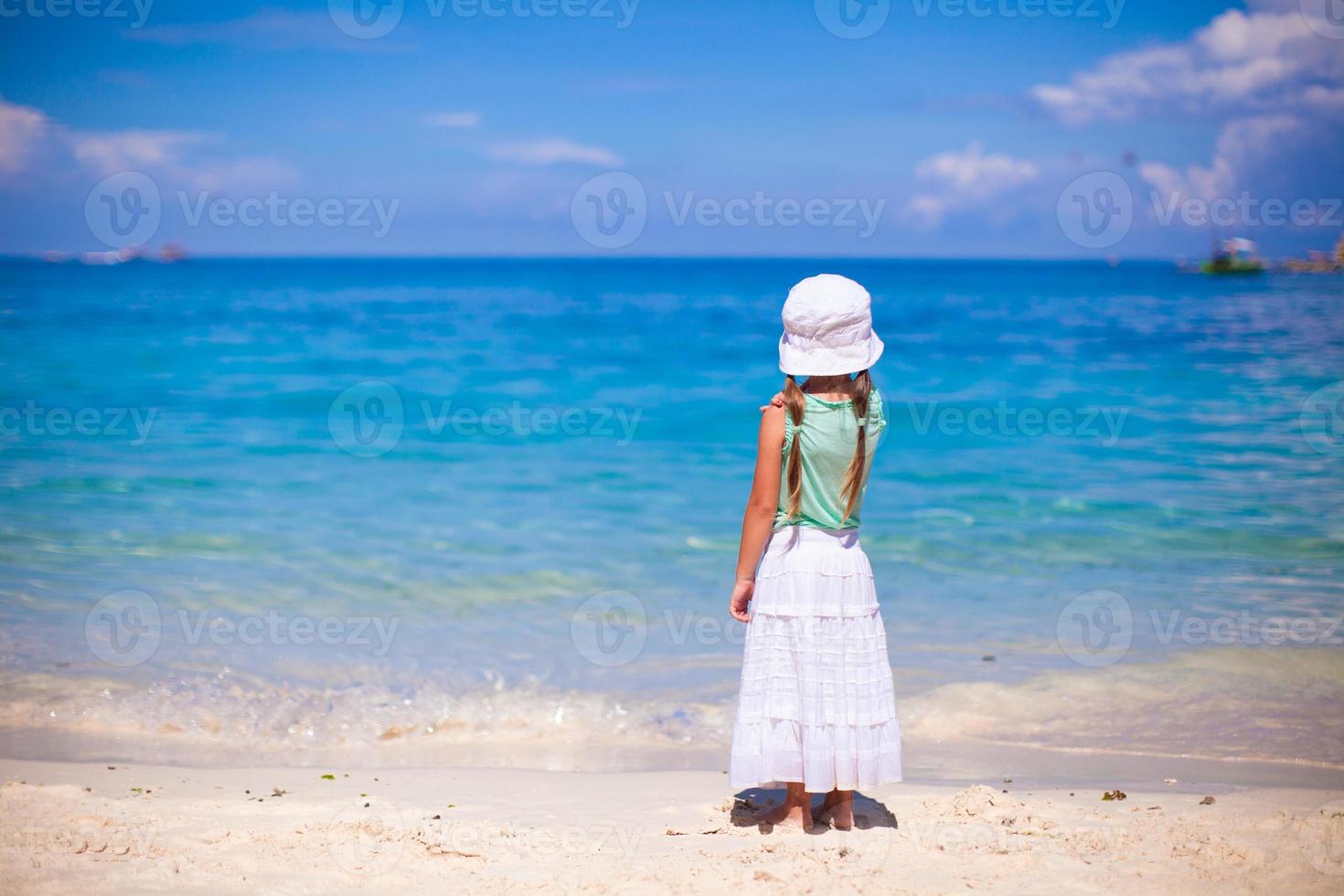 achterzijde visie van weinig meisje in hoed op zoek Bij de zee Aan wit zand strand foto