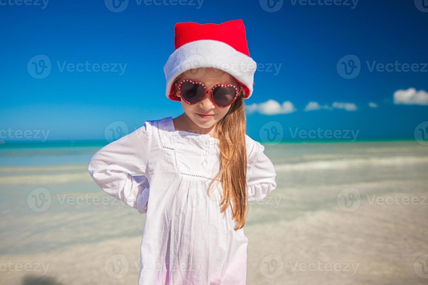 weinig meisje in rood hoed de kerstman claus en zonnebril Aan de exotisch strand foto