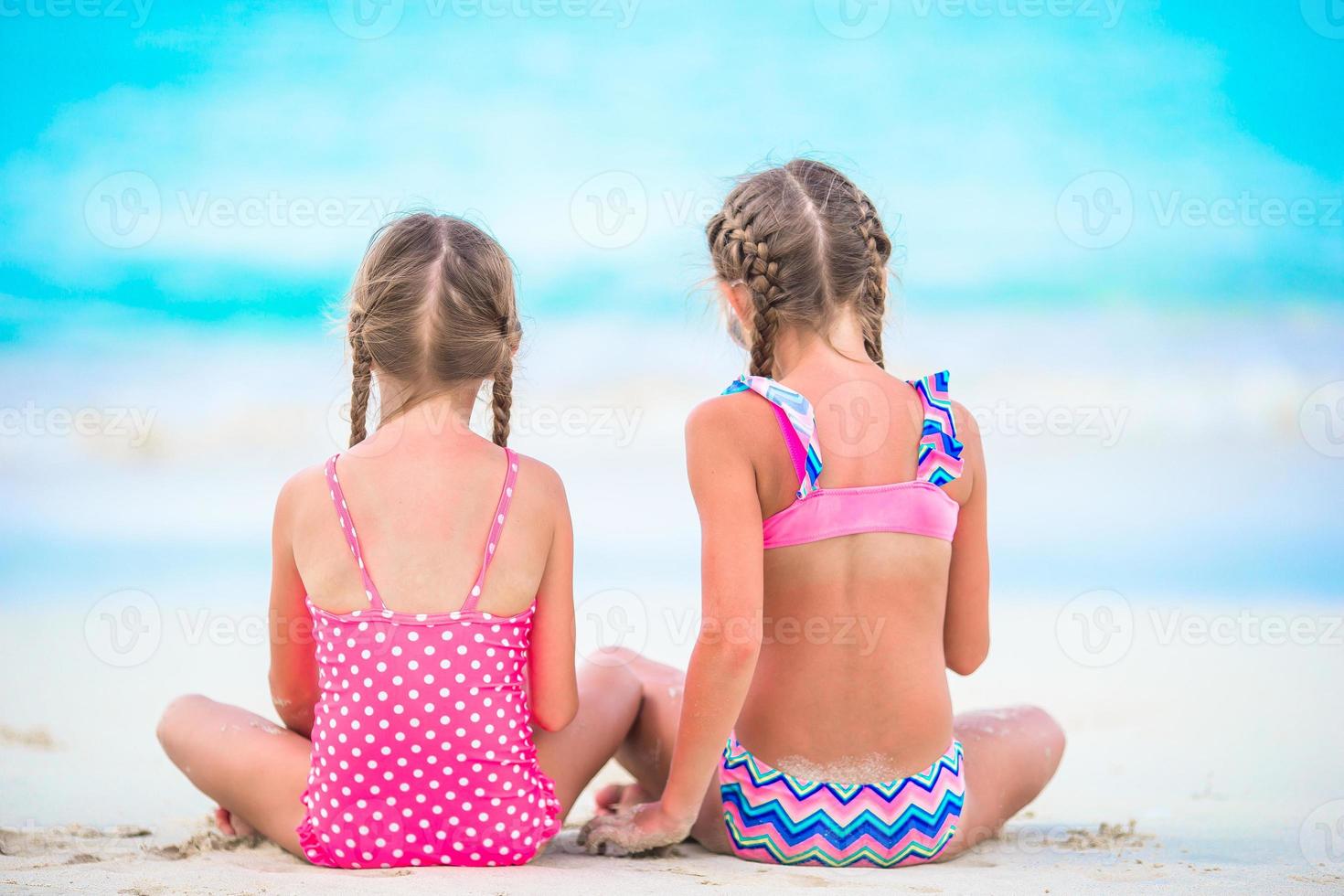 aanbiddelijk weinig meisjes spelen met zand Aan de strand. terug visie van kinderen zittend in Ondiep water en maken een Zandkasteel foto