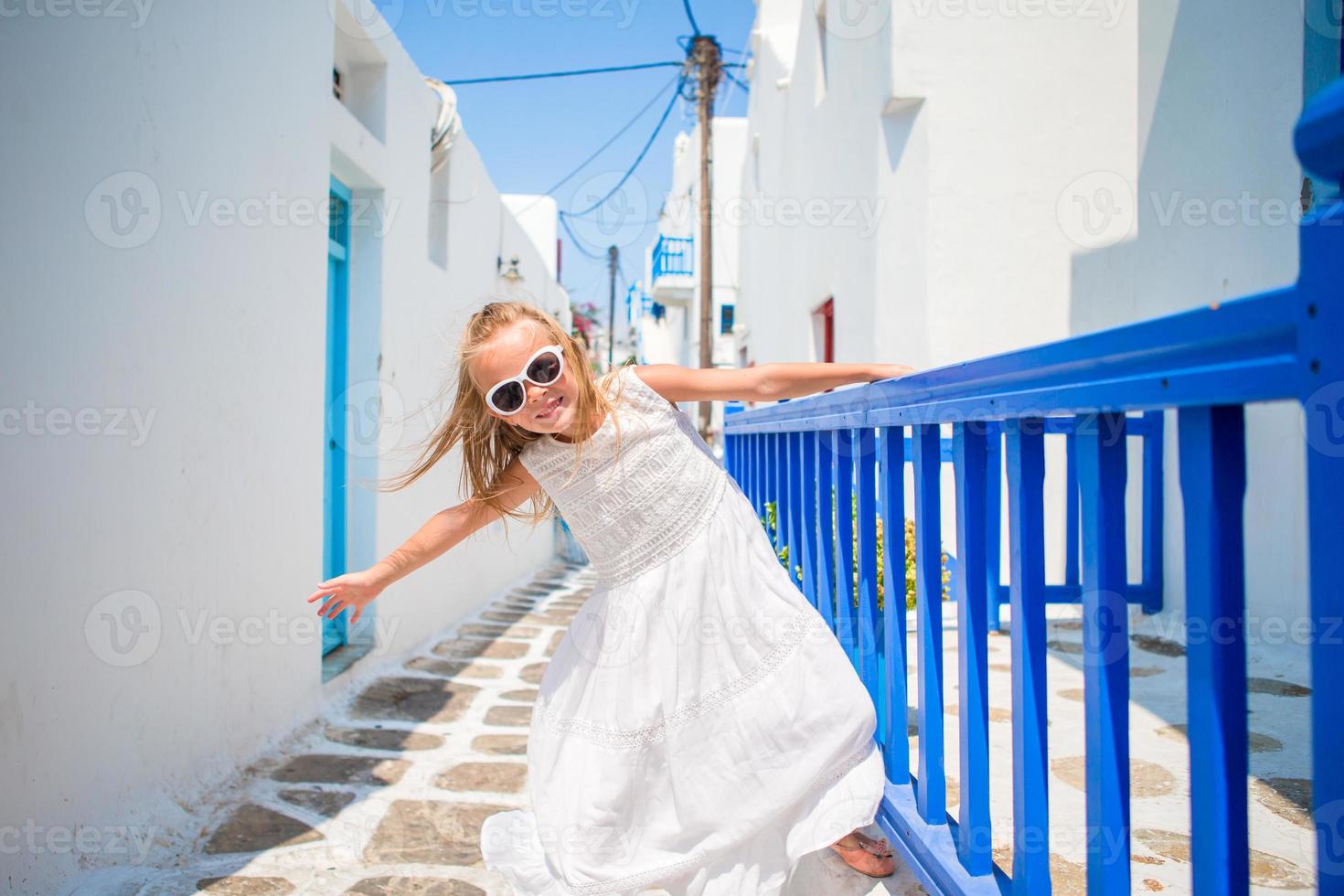 charmant meisje in wit jurk buitenshuis in oud straten een mykonos. kind Bij straat van typisch Grieks traditioneel dorp met wit muren en kleurrijk deuren Aan Mykonos eiland, in Griekenland foto