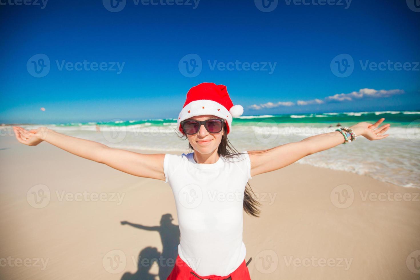 jong vrouw in de kerstman hoed wandelen verspreiding haar handen Aan wit zanderig strand foto